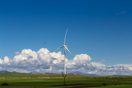A scenic Georgian landscape featuring towering wind turbines amidst lush green fields under a bright blue sky. Snow-capped mountains rise in the distance, framed by fluffy white clouds. A vision of clean energy and nature's harmonyの写真素材