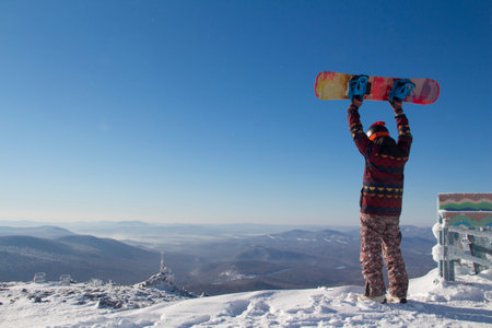 A man in snowboarding gear on the top of a mountain. Young man standing with a snowboard on the top...