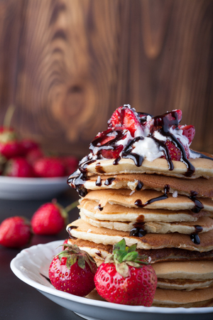 Stack of pancakes with strawberries, whip cream and chocolate syrup on a white plate on a wooden background. Vertical image. Copy space.の写真素材