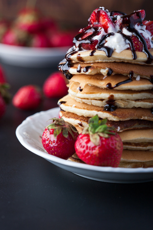 Stack of pancakes with strawberries, whip cream and chocolate syrup on a white plate on a black background. Vertical image. Copy space.の写真素材