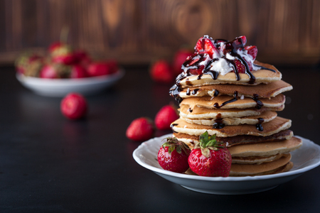 Stack of pancakes with strawberries, whip cream and chocolate syrup on a white plate on a wooden black background. Copy space.の写真素材