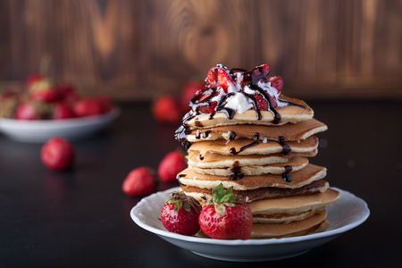 Stack of pancakes with strawberries, whip cream and chocolate syrup on a white plate on a wooden background. Copy space.の写真素材