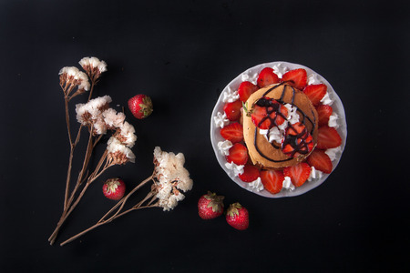 Homemade pancakes with strawberries, whipped cream and chocolate topping, decorated with flowers on black background. Overhead shot. Flat lay. Copy space.の写真素材