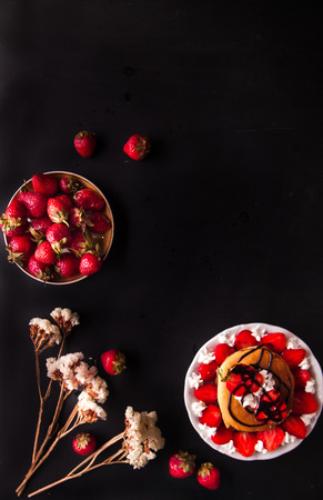 Homemade pancakes with strawberries, whipped cream and chocolate topping, decorated with flowers on black background. Overhead shot. Flat lay. Copy space. Vertical imageの写真素材
