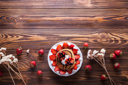 Homemade pancakes with strawberries, whipped cream and chocolate topping, decorated with flowers on wooden background. Overhead shot. Flat lay. Copy space.の写真素材
