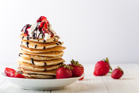 Stack of pancakes with strawberries, whip cream and chocolate syrup on a white plate on a white background. Copy space. Horizontal imageの写真素材