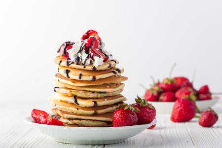 Stack of pancakes with strawberries, whip cream and chocolate syrup on a white plate on a white background. Copy space. Horizontal image.の写真素材