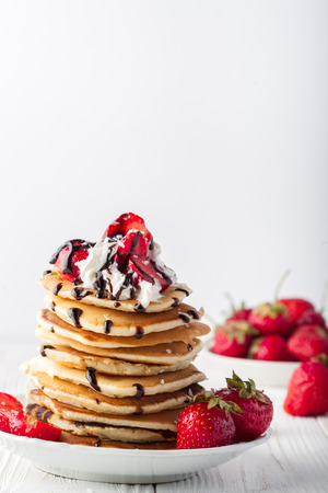 Stack of pancakes with strawberries, whip cream and chocolate syrup on a white plate on a white background. Copy space.の写真素材