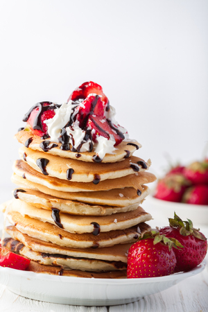 Stack of pancakes with strawberries, whip cream and chocolate syrup on a white plate on a white background. Copy space.の写真素材