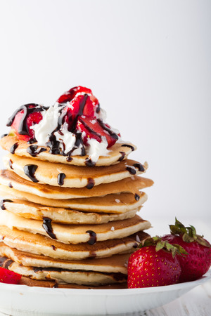 Stack of pancakes with strawberries, whip cream and chocolate syrup on a white plate on a white background. Copy space.の写真素材