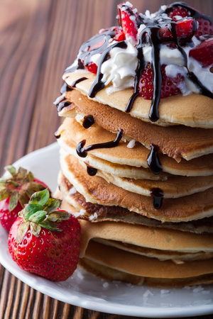 Stack of pancakes with strawberries, whip cream and chocolate syrup on a white plate on a wooden background. Vertical image. Copy space. Close up. Macroの写真素材