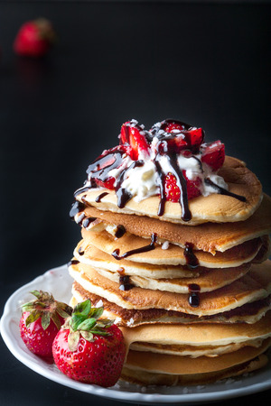 Stack of pancakes with strawberries, whip cream and chocolate syrup on a white plate on a black background. Vertical image. Copy space.の写真素材