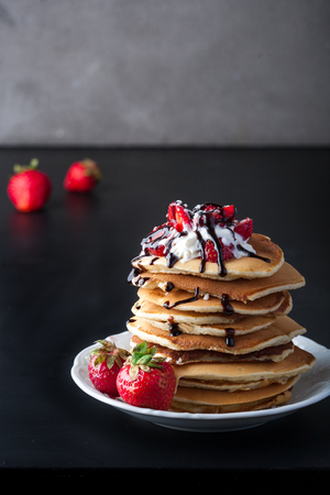Stack of pancakes with strawberries, whip cream and chocolate syrup on a white plate on a dark background. Vertical image. Copy space.の写真素材