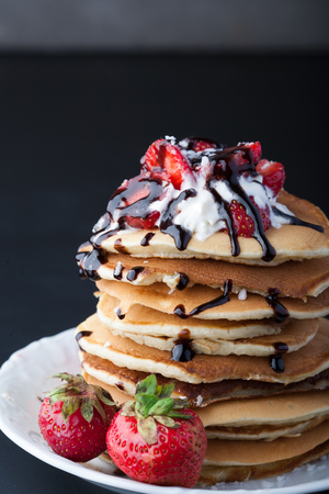 Stack of pancakes with strawberries, whip cream and chocolate syrup on a white plate on a black background. Vertical image. Copy space.の写真素材