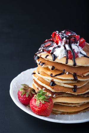Stack of pancakes with strawberries, whip cream and chocolate syrup on a white plate on a black background. Vertical image. Copy space.の写真素材
