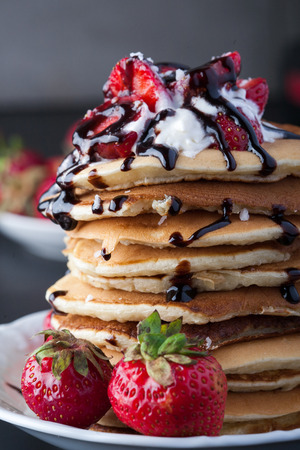 Stack of pancakes with strawberries, whip cream and chocolate syrup on a white plate on a black background. Vertical image. Copy space.の写真素材