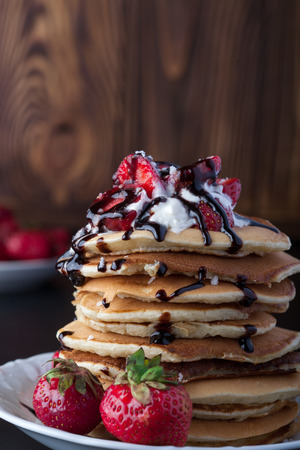 Stack of pancakes with strawberries, whip cream and chocolate syrup on a white plate on a wooden background. Vertical image. Copy space.の写真素材