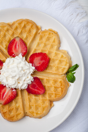 Norwegian heart shaped waffles topped with strawberries,  mint and whipped cream on white plate and white wooden background. St. Valentine's Day breakfast. Romantic breakfast. Copy spaceの写真素材