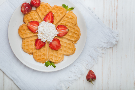 Norwegian heart shaped waffles topped with strawberries,  mint and whipped cream on white plate and white wooden background. St. Valentine's Day breakfast. Romantic breakfast. Copy spaceの写真素材