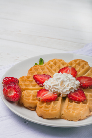 Norwegian heart shaped waffles topped with strawberries,  mint and whipped cream on white plate and white wooden background. St. Valentine's Day breakfast. Romantic breakfast. Copy spaceの写真素材