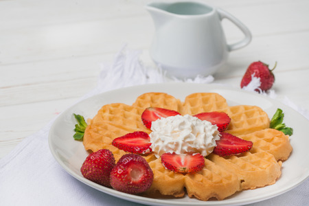 Norwegian heart shaped waffles topped with strawberries,  mint and whipped cream on white plate and white wooden background. St. Valentine's Day breakfast. Romantic breakfast. Copy spaceの写真素材
