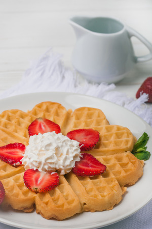 Norwegian heart shaped waffles topped with strawberries,  mint and whipped cream on white plate and white wooden background. St. Valentine's Day breakfast. Romantic breakfast. Copy spaceの写真素材