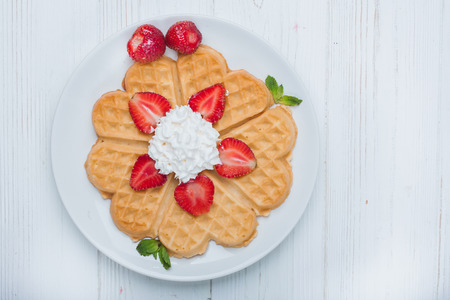 Norwegian heart shaped waffles topped with strawberries,  mint and whipped cream on white plate and white wooden background. St. Valentine's Day breakfast. Romantic breakfast. Copy spaceの写真素材