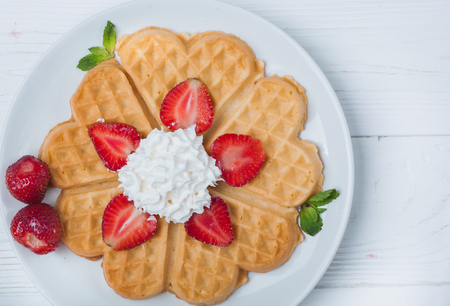 Norwegian heart shaped waffles topped with strawberries,  mint and whipped cream on white plate and white wooden background. St. Valentine's Day breakfast. Romantic breakfast. Copy spaceの写真素材
