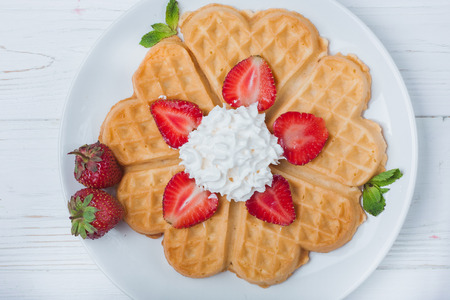 Norwegian heart shaped waffles topped with strawberries,  mint and whipped cream on white plate and white wooden background. St. Valentine's Day breakfast. Romantic breakfast. Copy spaceの写真素材