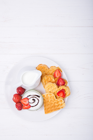 Breakfast scene. Heart shaped waffles with strawberries, cream sauce with white pitcher with milk on white plate and white wooden background. St Valentines day breakfast. Romantic. Copy spaceの写真素材