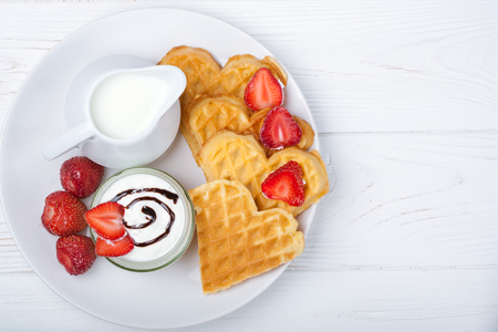 Breakfast scene. Heart shaped waffles with strawberries, cream sauce with white pitcher with milk on white plate and white wooden background. St Valentines day breakfast. Romantic. Copy spaceの写真素材