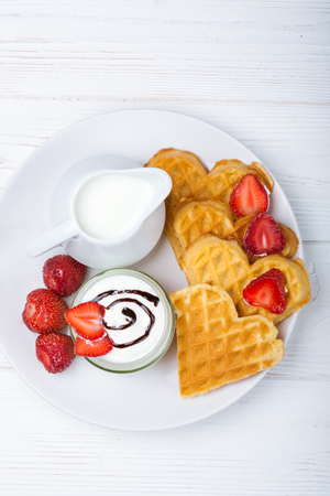 Breakfast scene. Heart shaped waffles with strawberries, cream sauce with white pitcher with milk on white plate and white wooden background. St Valentines day breakfast. Romantic. Copy spaceの写真素材