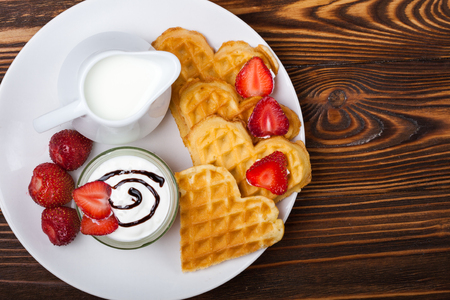 Heart shaped waffles with strawberries, cream sauce, pitcher with milk on white plate and wooden background. St Valentines day breakfast. Romantic breakfast dessert. Copy spaceの写真素材