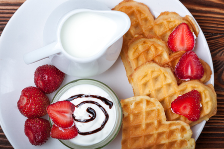Heart shaped waffles with strawberries, cream sauce, pitcher with milk on white plate and wooden background. St Valentines day breakfast. Romantic breakfast dessert. Copy spaceの写真素材