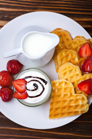 Heart shaped waffles with strawberries, cream sauce, pitcher with milk on white plate and wooden background. St Valentines day breakfast. Romantic breakfast dessert. Copy spaceの写真素材