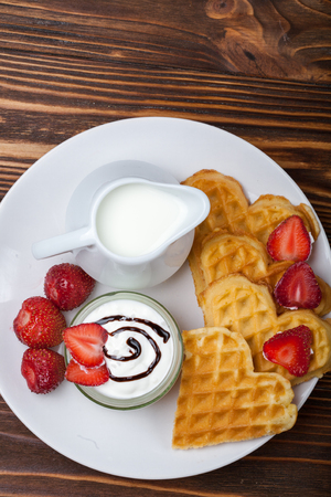 Heart shaped waffles with strawberries, cream sauce, pitcher with milk on white plate and wooden background. St Valentines day breakfast. Romantic breakfast dessert. Copy spaceの写真素材