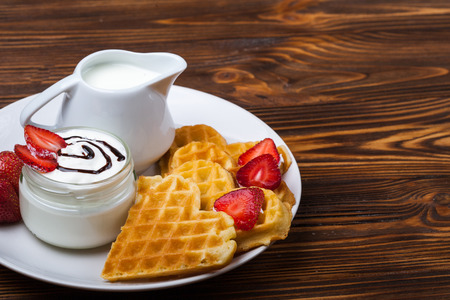 Heart shaped waffles with strawberries, cream sauce, pitcher with milk on white plate and wooden background. St Valentines day breakfast. Romantic breakfast dessert. Copy spaceの写真素材