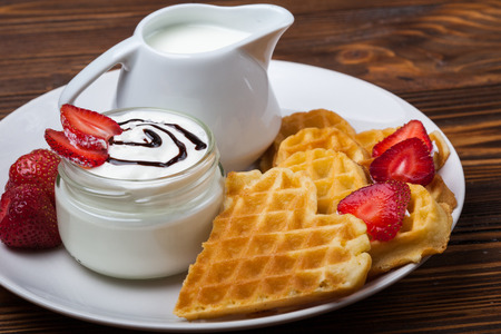 Heart shaped waffles with strawberries, cream sauce, pitcher with milk on white plate and wooden background. St Valentines day breakfast. Romantic breakfast dessert. Copy spaceの写真素材