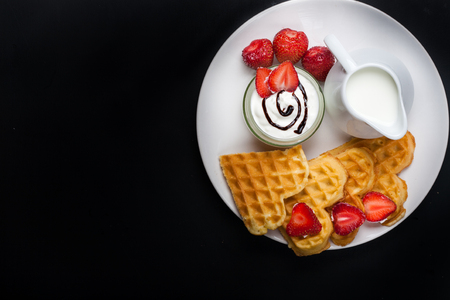 Breakfast scene. Heart shaped waffles with strawberries, cream sauce, pitcher with milk on white plate and black background. St Valentines day breakfast. Romantic breakfast dessert. Copy spaceの写真素材