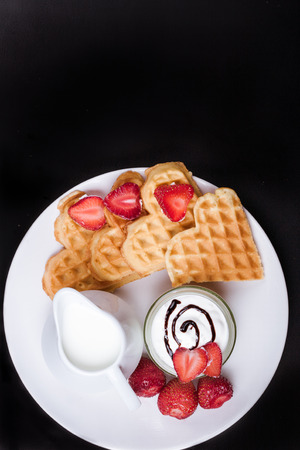 Breakfast scene. Heart shaped waffles with strawberries, cream sauce, pitcher with milk on white plate and black background. St Valentines day breakfast. Romantic breakfast dessert. Copy spaceの写真素材
