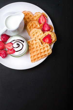 Breakfast scene. Heart shaped waffles with strawberries, cream sauce, pitcher with milk on white plate and black background. St Valentines day breakfast. Romantic breakfast dessert. Copy spaceの写真素材