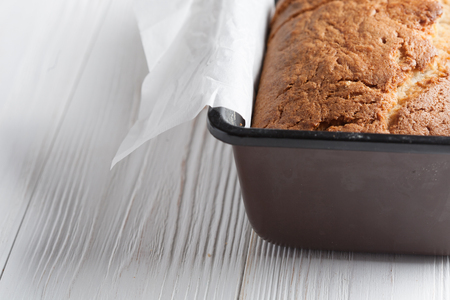 Banana bread in a pan with white parchment paper on a white wooden table. Close up. Bread cracks pattern. Bakery conceptの写真素材
