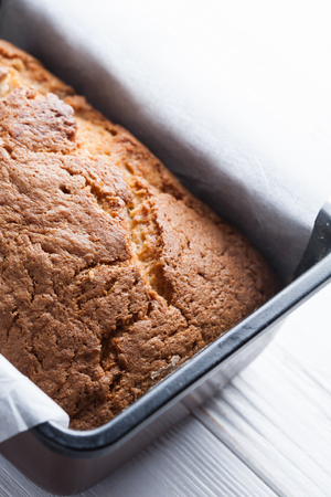 Banana bread in a pan with white parchment paper on a white wooden table. Close up. Bread cracks pattern. Bakery conceptの写真素材
