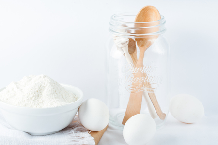Baking ingredients. White background. Ingredients and tools to make a cake. Flour, milk, eggs, rolling pin, wooden spoons, jar. Copy space. Bakery. Baking conceptの写真素材