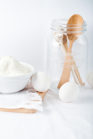 Baking ingredients. White background. Ingredients and tools to make a cake. Flour, milk, eggs, rolling pin, wooden spoons, jar. Copy space. Bakery. Baking conceptの写真素材