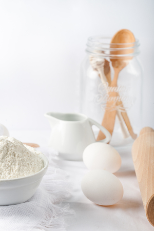 Baking ingredients. White background. Ingredients and tools to make a cake. Flour, milk, eggs, rolling pin, wooden spoons, jar. Copy space. Bakery. Baking conceptの写真素材