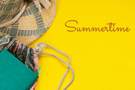 Straw hat, notepad, backpack and Summertime text on bright yellow background with copy space. Summer...