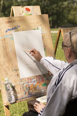 Mature grey-haired woman draws floral sketch outdoors in art studio in sunny day. Moscow - June 1, 2019. Hobby for retired people, social programs for elderly people. Selective focusのeditorial素材