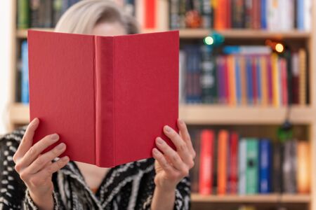 Woman sitting on a floor and holding book in front of face on blurred bookshelf backgroundの写真素材