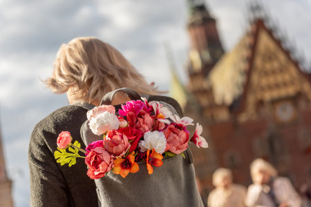 Photo from the back of young blonde romantic woman with bouquet of flowers in grey backpack,...